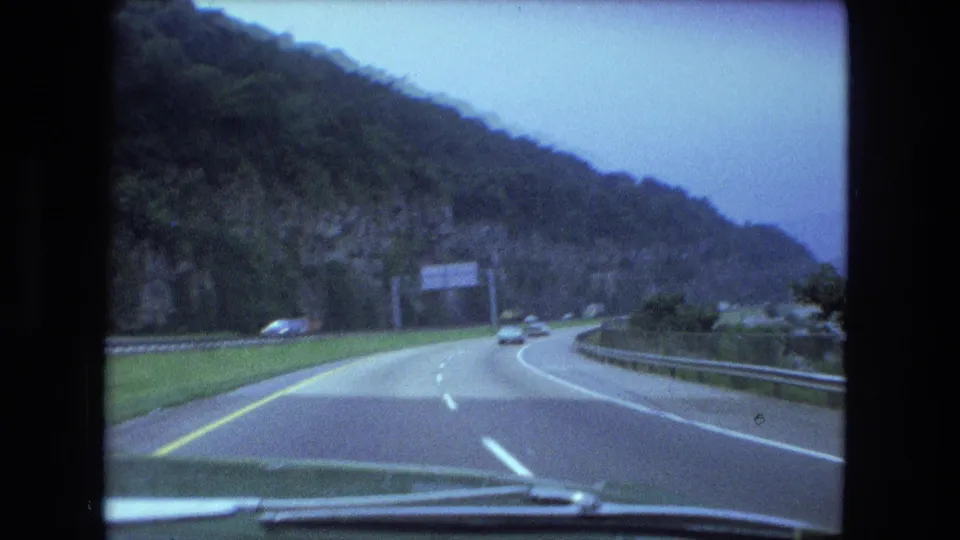 Windshield perspective on a curving highway, with a wooded rock face, center lane markings, guardrails, and cars in the distance.