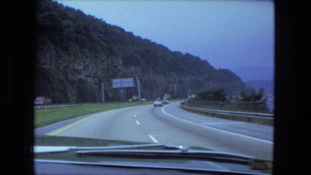 1974 8mm windshield view of a multi-lane highway curving past a wooded rock face with cars ahead and an overhead sign.