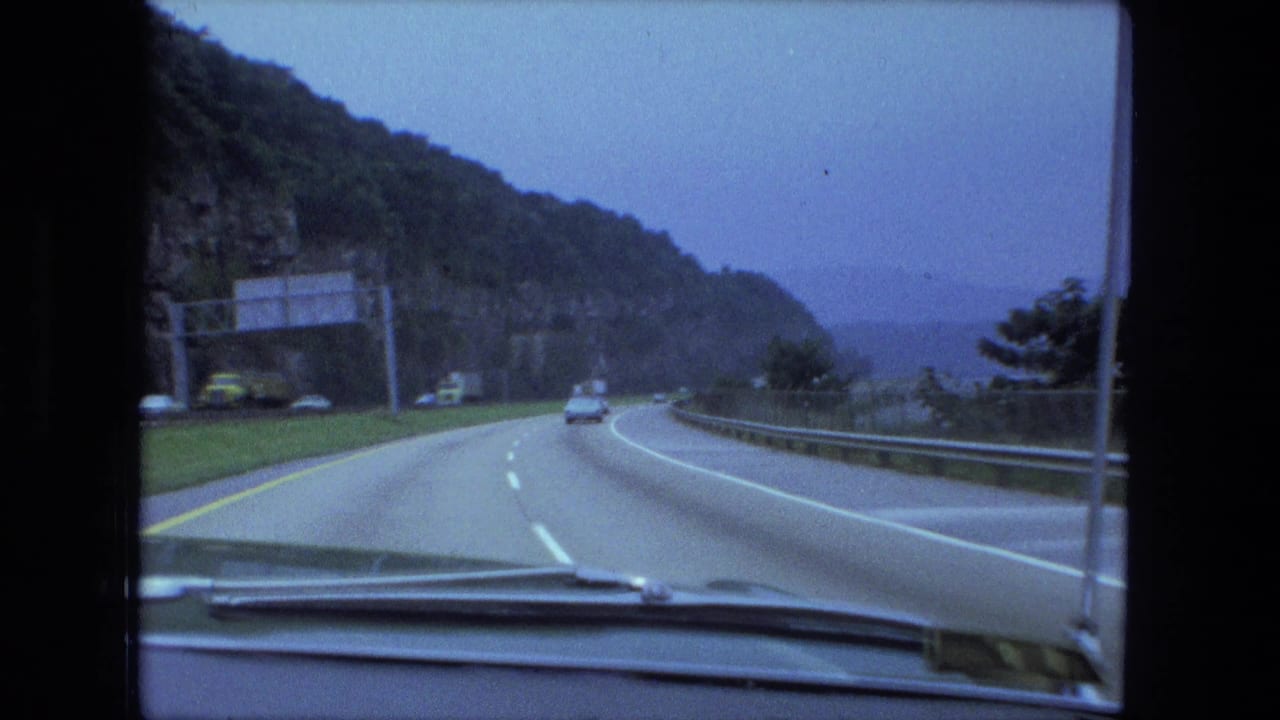Windshield view from 1974 home-movie footage showing a divided highway, classic-era cars, rock slope, and signs ahead.