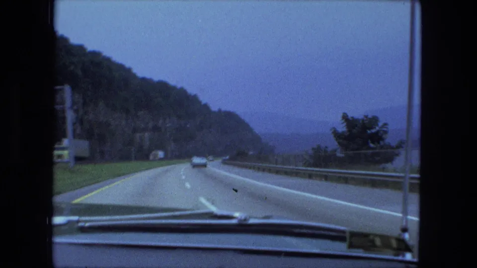 1974 8mm car-window perspective of a divided highway following a wooded slope, with distant haze or water beyond the guardrail.