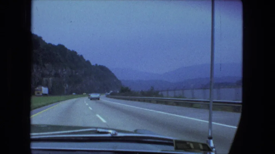 Late clip frame shows a windshield view of the highway beside a wooded rock face, with guardrail, traffic, and distant water or haze.