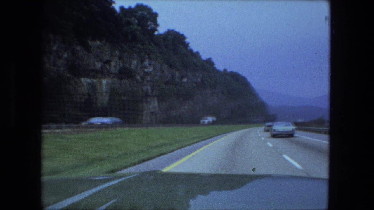 Two-lane highway beside a rocky cliff with cars ahead under an overcast sky.