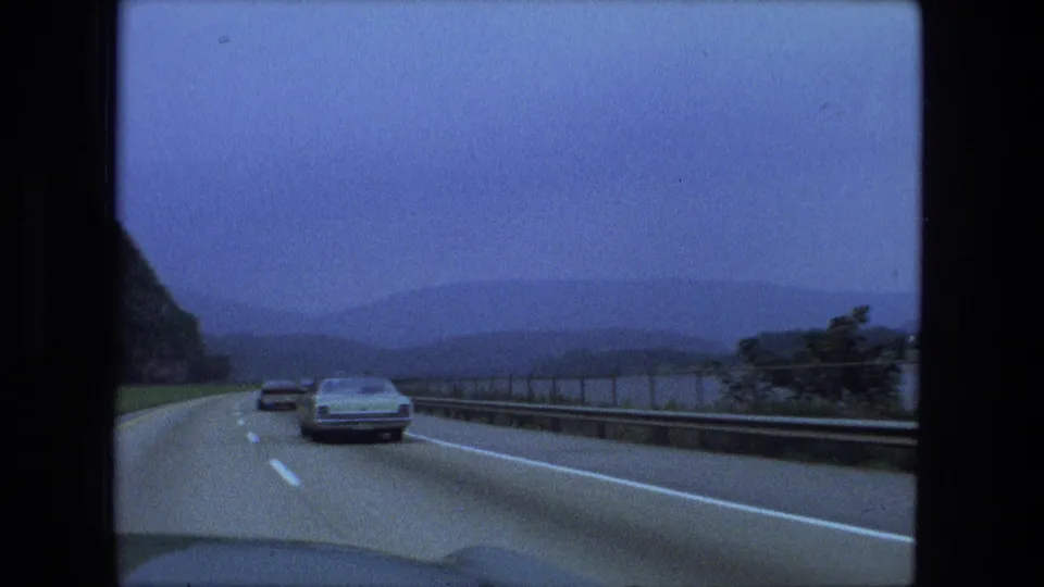 1974 8mm frame from a moving car, looking down a highway toward vehicles, guardrail, lake water, and hazy hills.