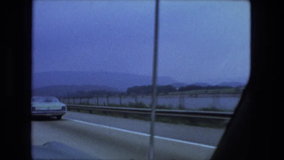 1974 8mm home-movie road view showing a car ahead, roadside guardrail, lake water, and muted hills beyond the highway.