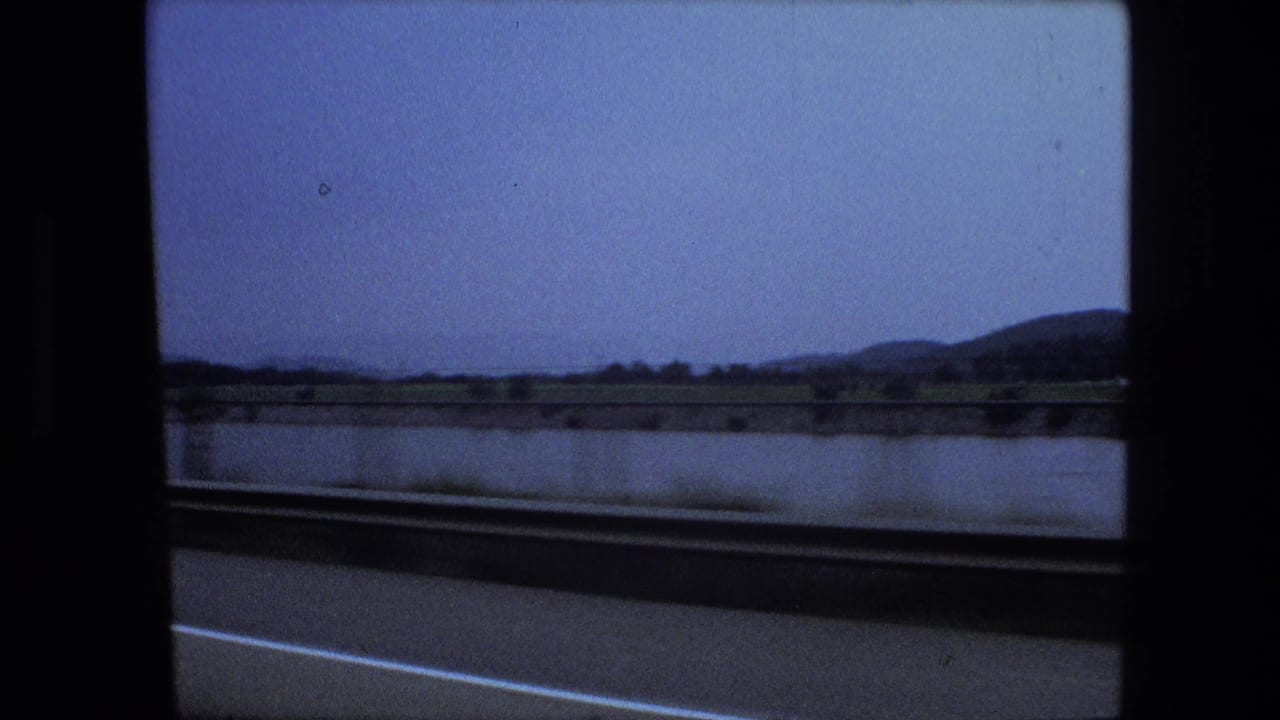 View through a vehicle window of road, water, distant low hills, and cloudy sky.