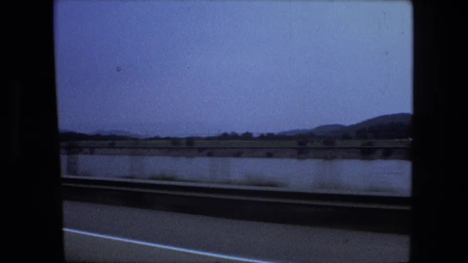 1974 8mm home-movie frame showing a passing road view beside water, with low hills in the distance and the car window frame visible.