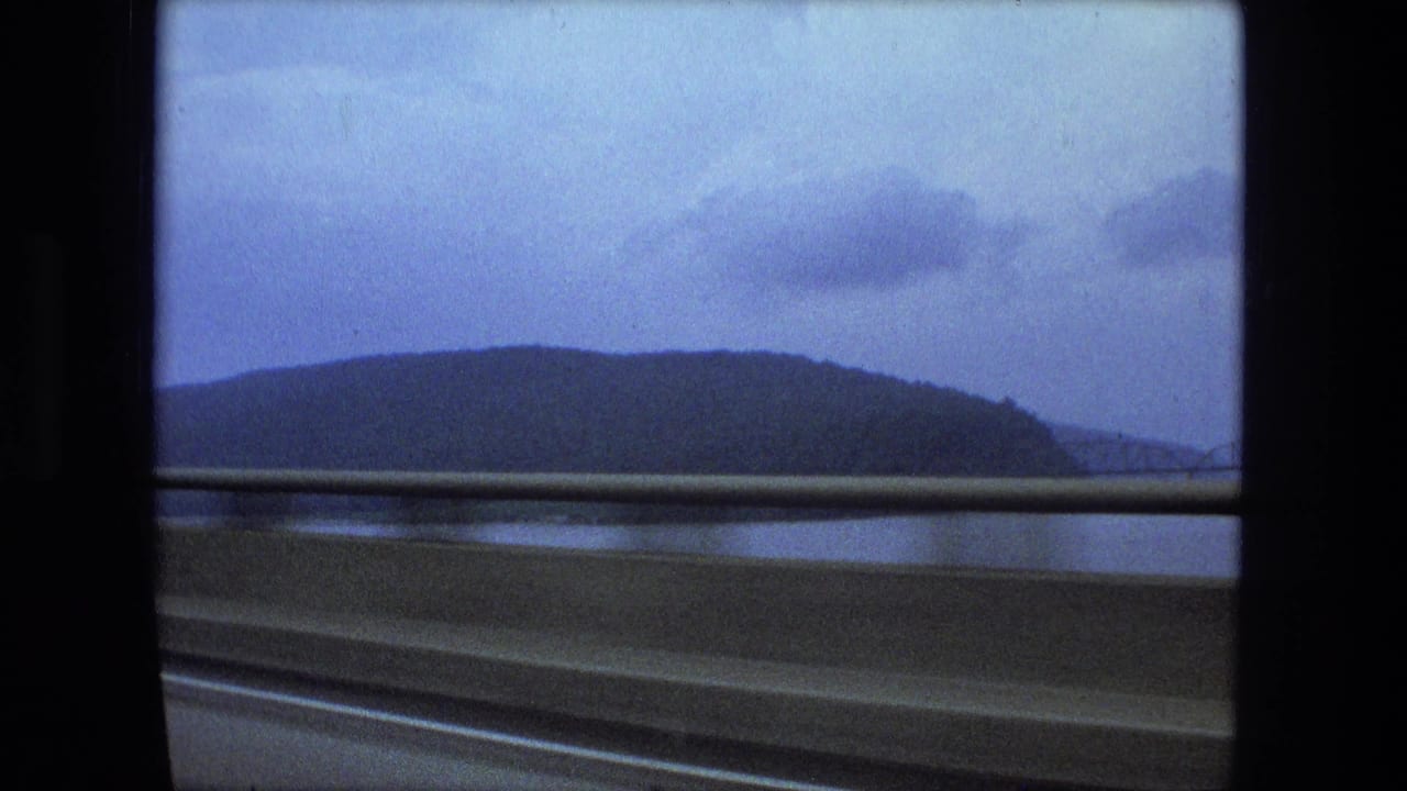 Guardrail, water, wooded hill, clouds, and window frame seen from a moving vehicle.