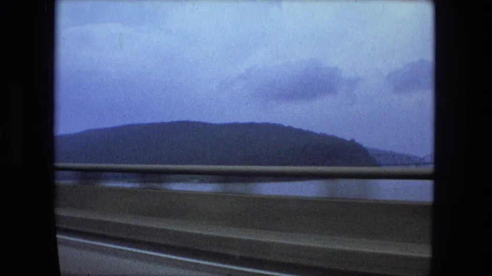 Passenger-side 8mm view from the 1974 clip, with guardrail in front of water and a dark hill beneath a cloudy blue sky.