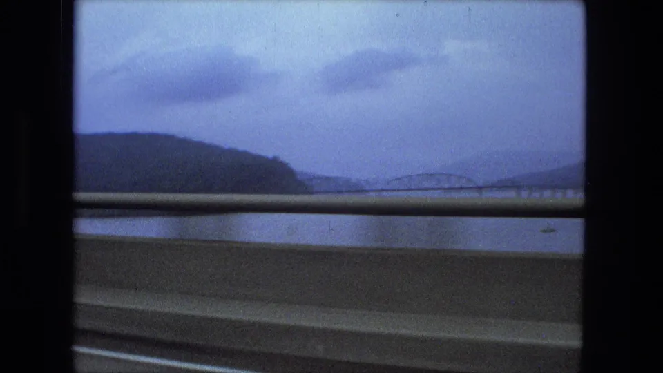 1974 8mm travel frame showing a distant arched bridge across water, filmed past a highway guardrail from inside a moving vehicle.