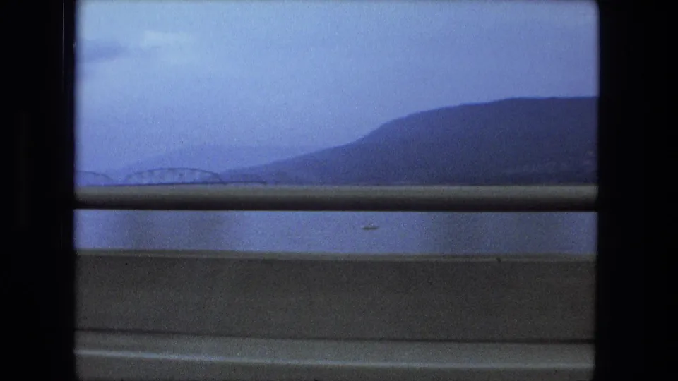 Final 1974 home-movie frame with a large hill beyond water, a faint bridge span, and a highway guardrail in the foreground.