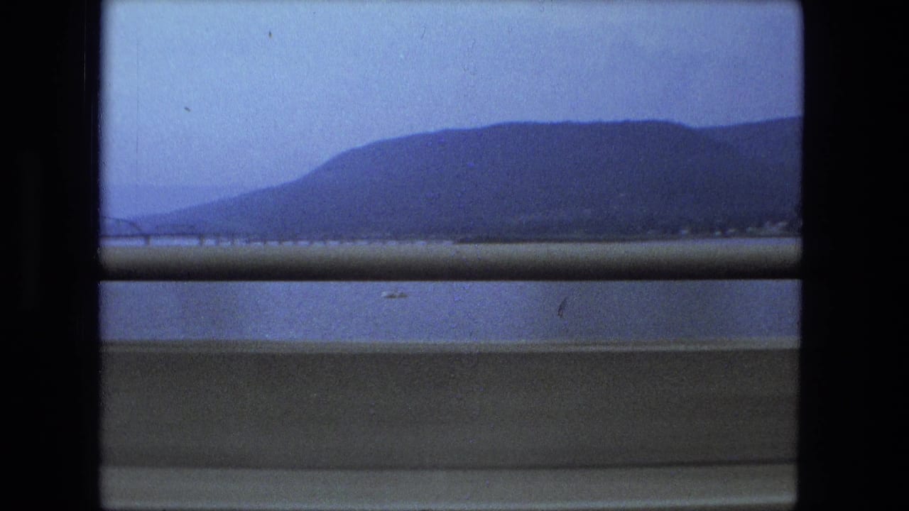Water and distant hills seen past a roadside barrier from a moving car.