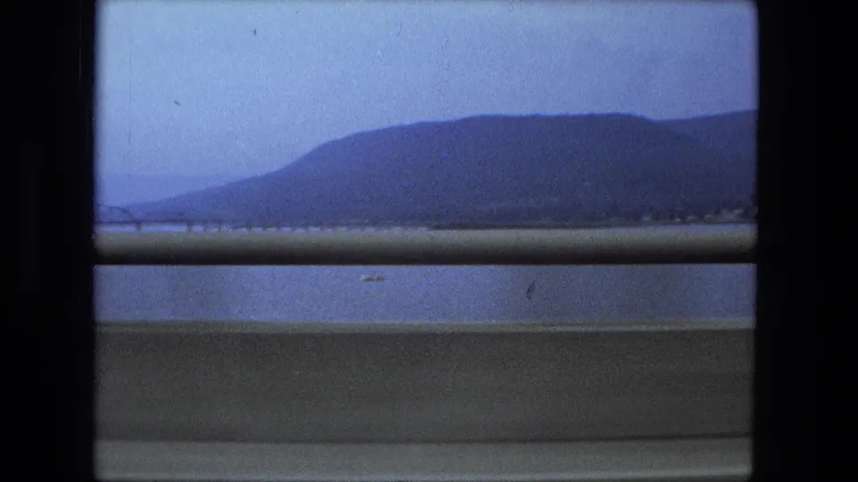 1974 8mm travel view labeled Fort Wayne, Indiana, showing water, distant hills, and a concrete roadside barrier from a moving vehicle.