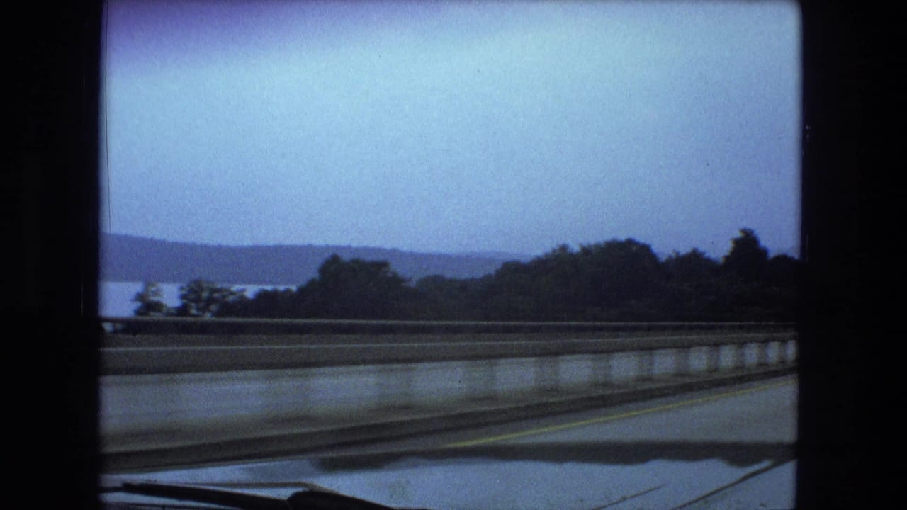Highway barrier, car hood, trees, water, and hills seen from inside a car.