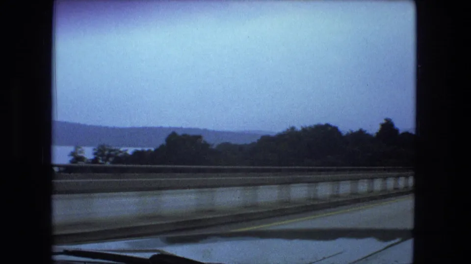 Moving-car view on 8mm film with a highway barrier, car hood, wooded shoreline, water, and low hills under a blue sky.