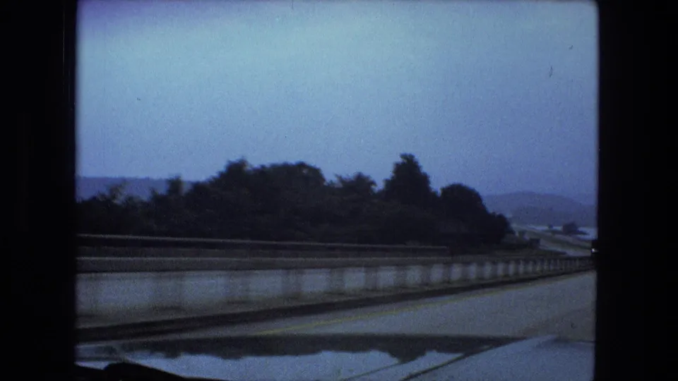 Passenger-side travel view showing a concrete barrier, dark tree line, water, and distant hills in soft 8mm color.