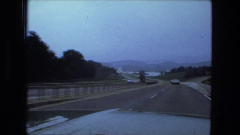 Forward-facing 1974 8mm driving view labeled Fort Wayne, Indiana, with highway lanes, vehicles, barrier, water, trees, and distant hills.