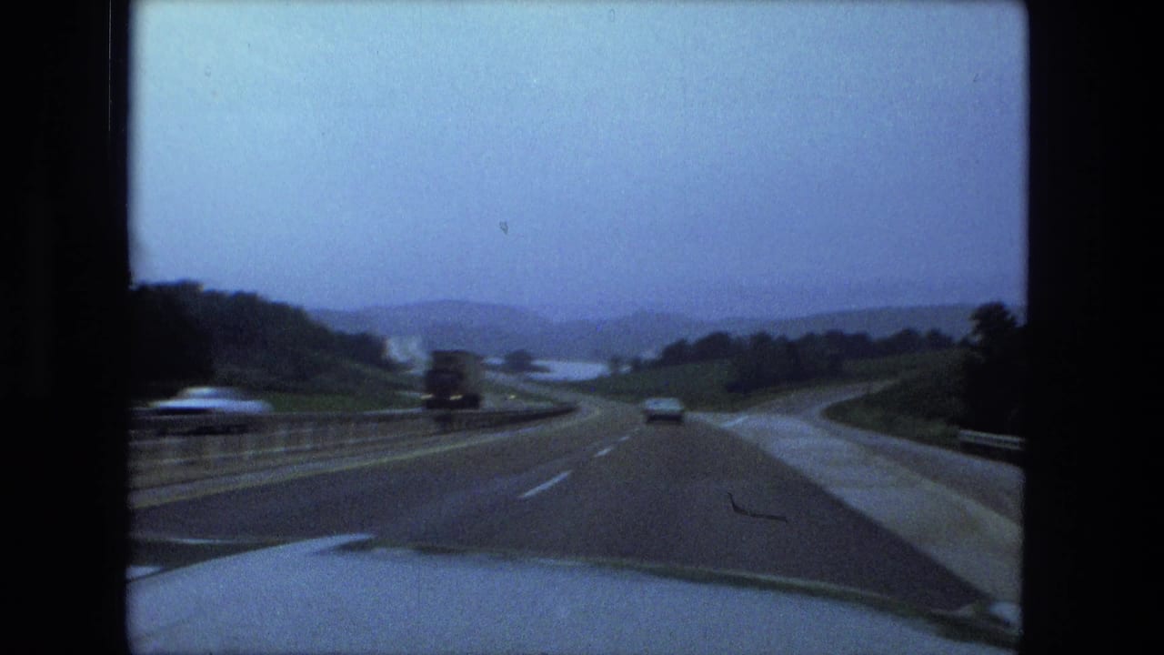 Highway seen through a vehicle window with cars ahead, a guardrail, wet pavement, and distant hills.