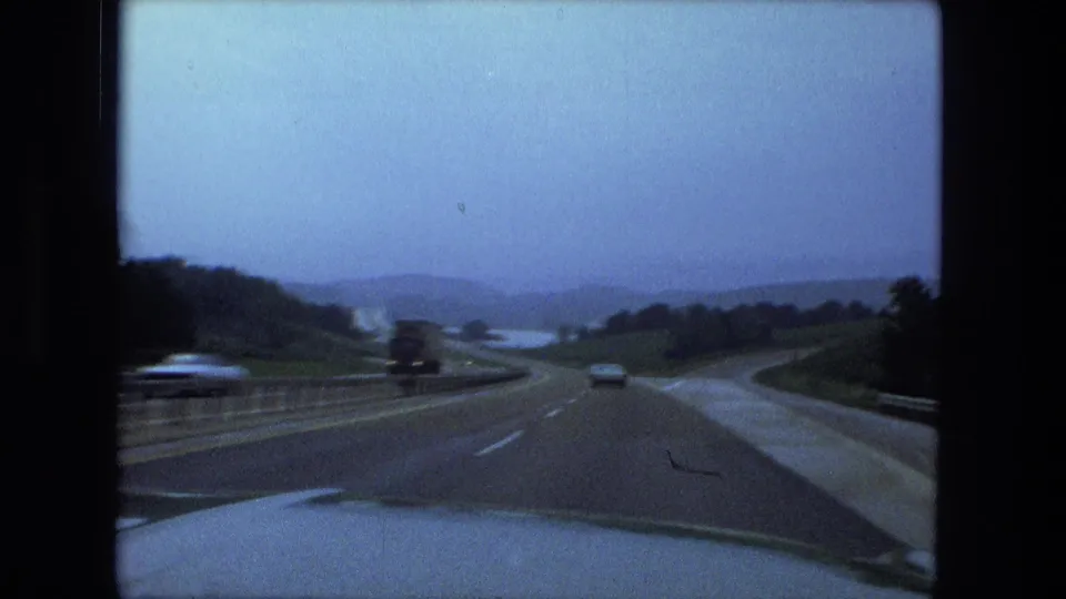 1974 Fort Wayne-area road footage showing a highway from inside a vehicle, with cars ahead, a guardrail, wet pavement, and distant hills.