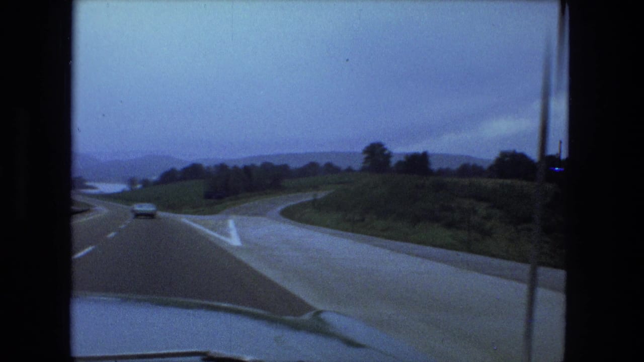 1974 Fort Wayne-area in-car view of a curving highway beside an exit ramp, grassy roadside, wet pavement, and distant water.