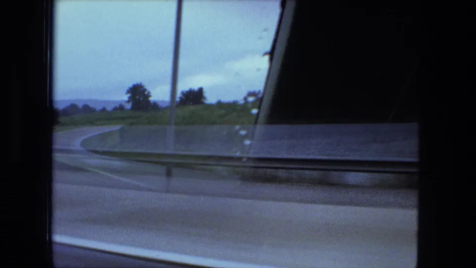 1974 Fort Wayne-area side-window view from a moving vehicle, showing a road curve, roadside pole, grassy verge, and car door frame.