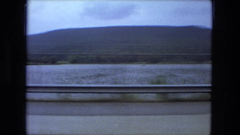 1974 Fort Wayne-area side-window view across wet pavement and water toward low hills beneath an overcast sky.