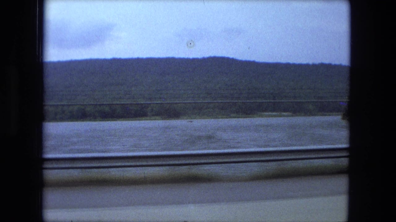1974 8mm view near Fort Wayne, Indiana, showing a river and low wooded hill through a train window.