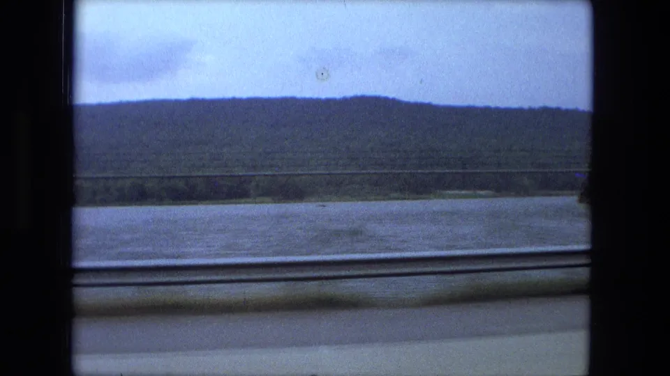 1974 8mm view near Fort Wayne, Indiana, showing a river and low wooded hill through a train window.