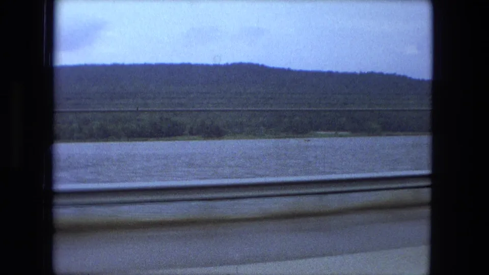 Passenger view from a moving train in 1974, with a broad river, dark wooded ridge, and overcast sky.