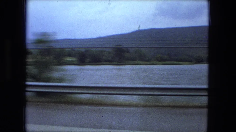 1974 home-movie frame with motion-blurred shoreline, river water, and wooded hills beyond the train window.
