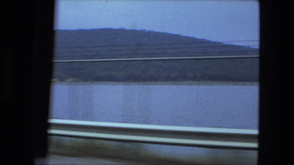 A 1974 8mm train-window view near Fort Wayne with river water, rounded wooded hillside, and pale overcast sky.