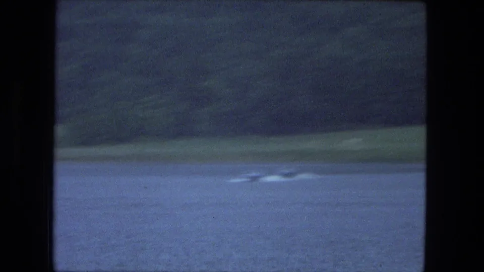 Moving-vehicle view of a lake and shoreline, with the boat wake faintly visible in 1974 8mm Fort Wayne-area footage.