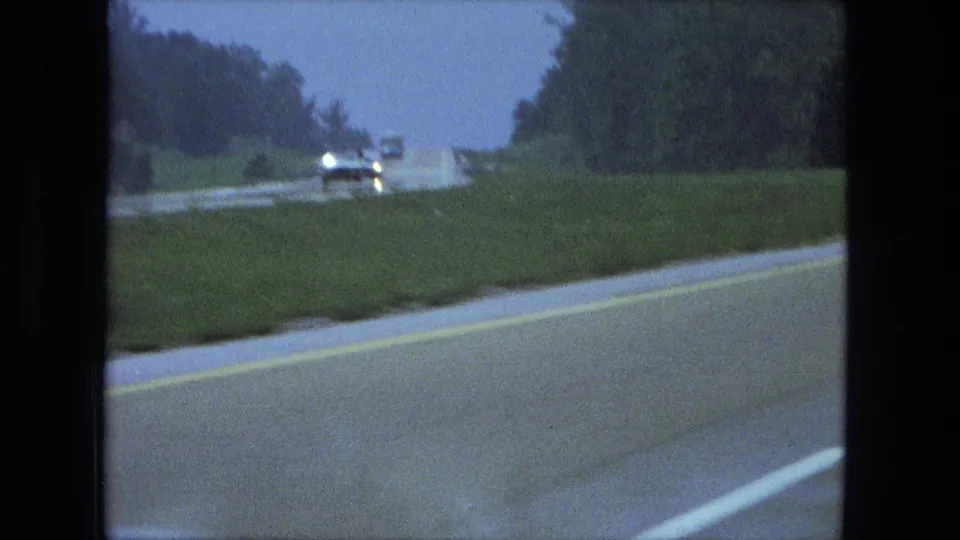 Rainy 1974 Fort Wayne road view with an approaching vehicle, headlamps, grass shoulder, and tree line.
