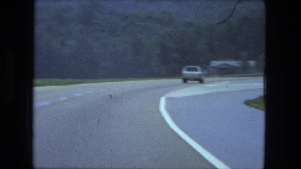 1974 Fort Wayne highway curve with a car traveling ahead on wet pavement beside grass and trees.