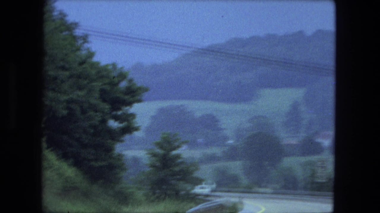 Curving two-lane road seen through trees with hills and fields in the distance.