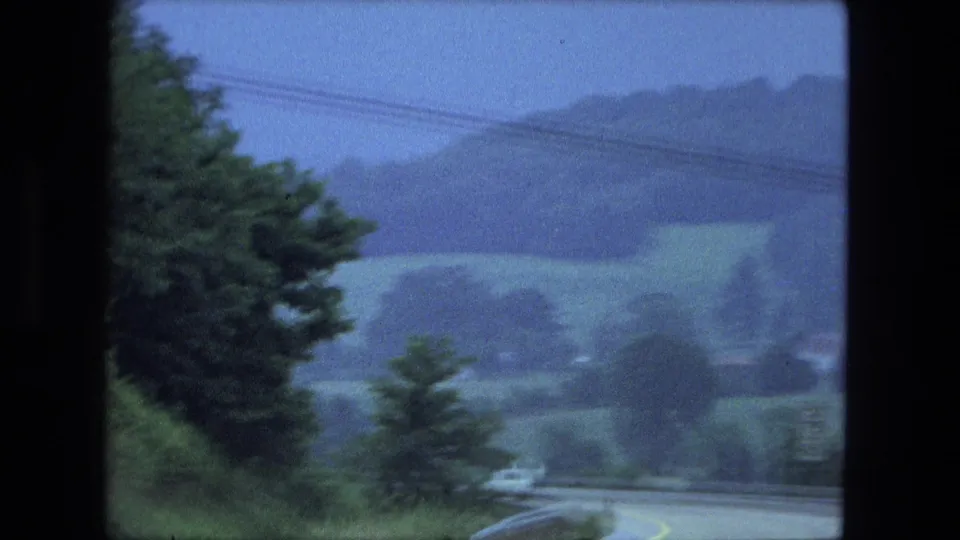 1974 8mm home-movie view from a moving vehicle on a curving rural road, with trees, utility lines, hills, and a pale car ahead.