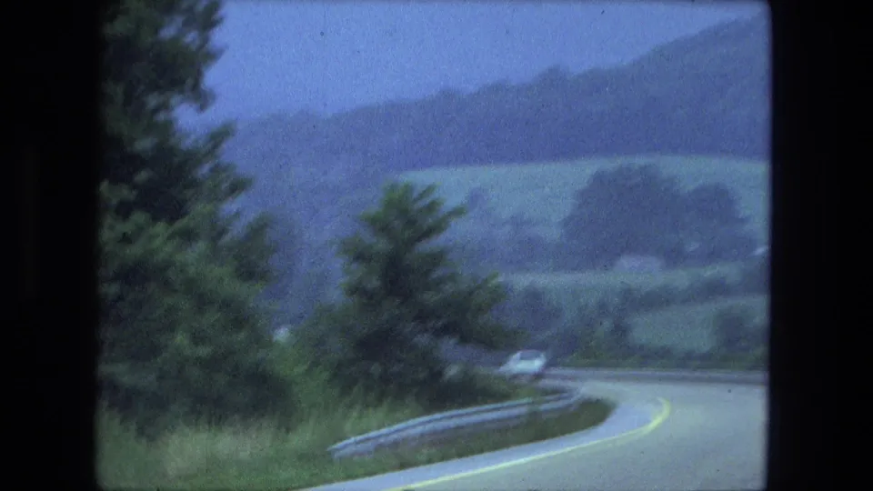 Moving-vehicle view of a winding rural road, guardrail, trees, and distant hills in blue-tinted 1970s 8mm footage.