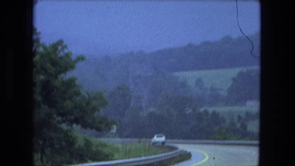 1974 home-movie frame showing a white car ahead on a downhill curve, with roadside trees, guardrail, fields, and hazy hills beyond.