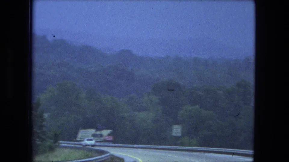 Blue-tinted 8mm frame of vehicles following a road curve past metal guardrails, roadside signs, trees, and wooded hills.