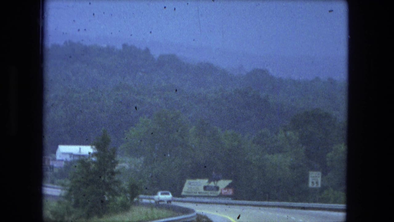Curving road through wooded hills with cars and a roadside speed sign.