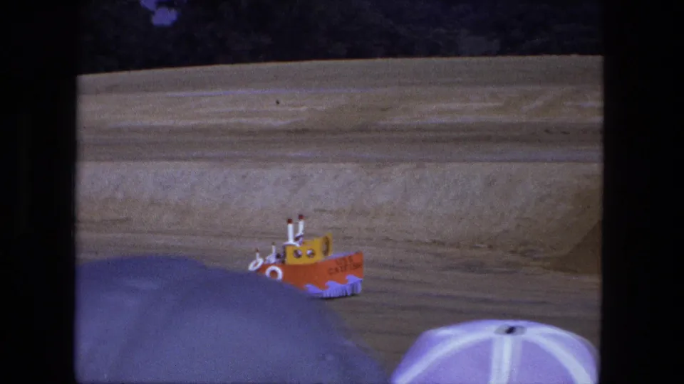 The USS CATFISH novelty boat vehicle continues across the sandy field, partly blocked by a purple and white umbrella in the foreground.