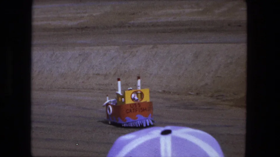 Rear view of the USS CATFISH boat-shaped vehicle on sand, with a purple and white umbrella in the foreground.