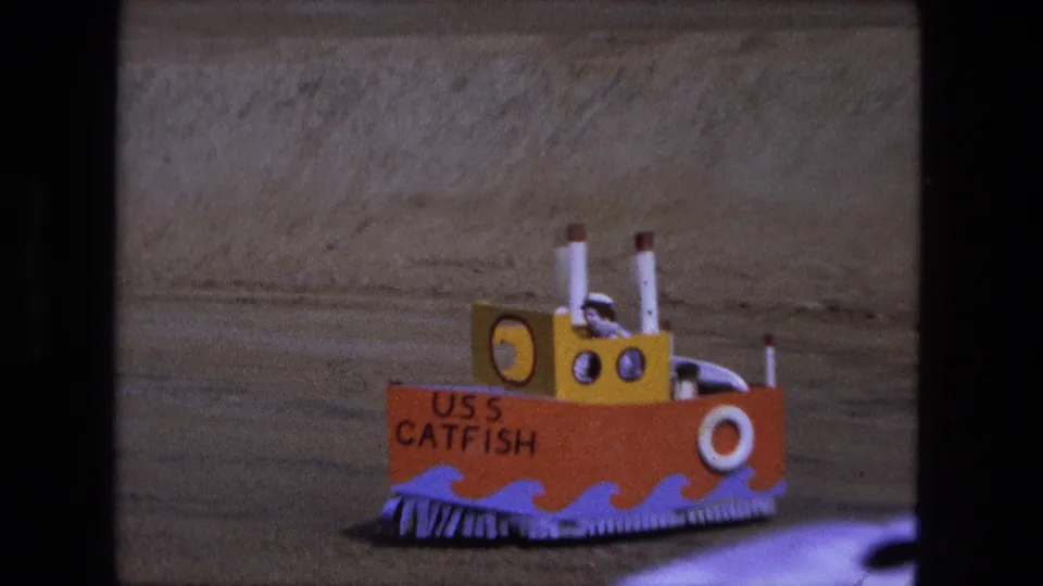 1974 Fort Wayne 8mm frame showing a colorful handmade USS Catfish model boat on sandy ground.