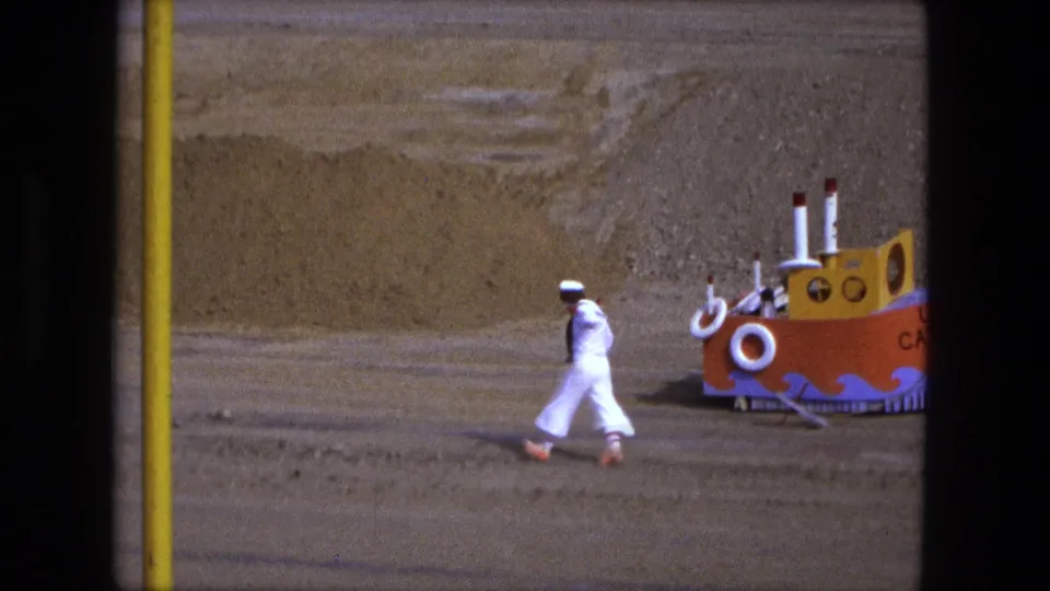 A person in a white sailor-style outfit runs across sandy ground near the handmade USS Catfish model in Fort Wayne, 1974.