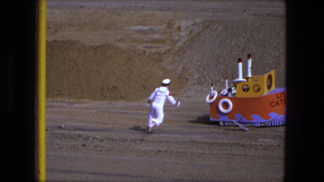 Child in a white sailor suit runs toward a colorful toy boat on sandy ground.