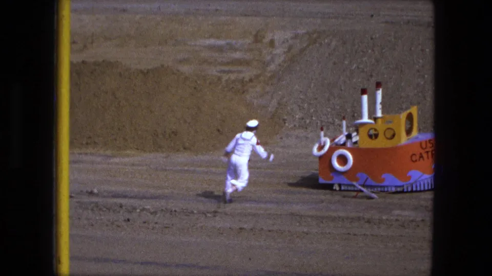 In Fort Wayne, Indiana, 1974 home-movie footage, a child in a white sailor suit approaches a handmade orange and yellow boat labeled USS Catfish.