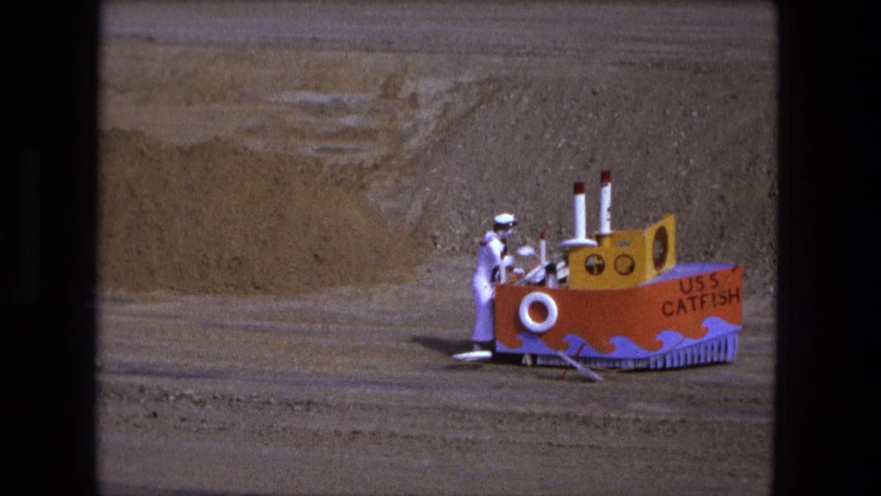 The child in a sailor costume stands next to the bright handmade USS Catfish boat, with sandy ground and dirt bank behind it.