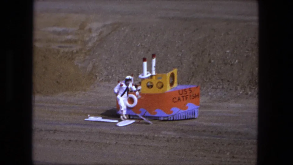 The child remains beside the orange USS Catfish prop boat, with pale boards set out on the sandy lot in Fort Wayne, Indiana, 1974 footage.