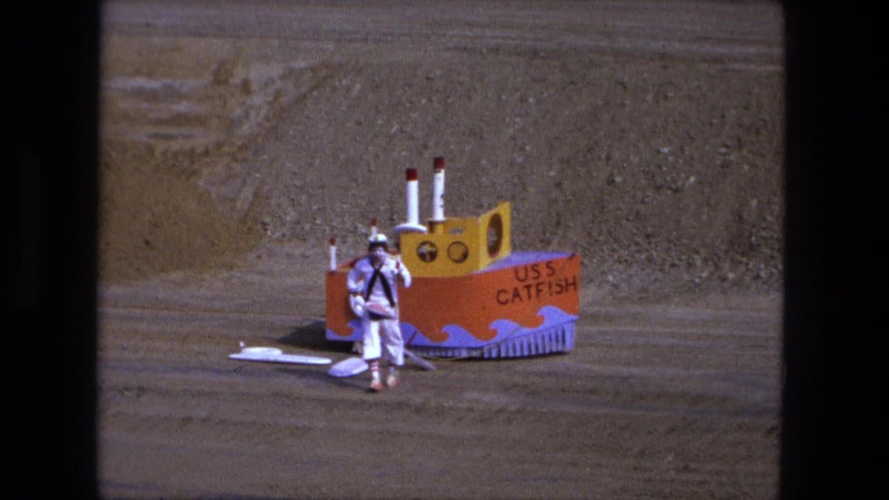 Person in a white sailor costume beside a colorful toy boat on a dirt field.