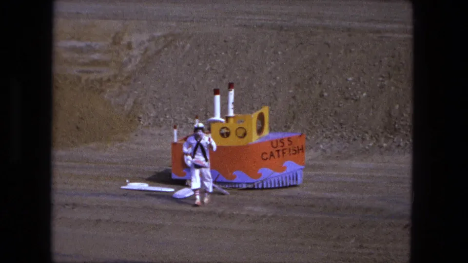 1974 Fort Wayne 8mm frame showing a costumed sailor figure near a handmade boat labeled USS CATFISH in an open dirt area.