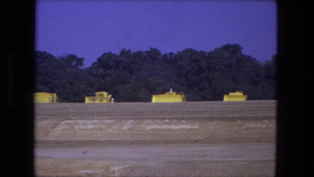 Wide 1974 Fort Wayne home-movie view of yellow construction vehicles lined up beyond a bare dirt field and tree line.